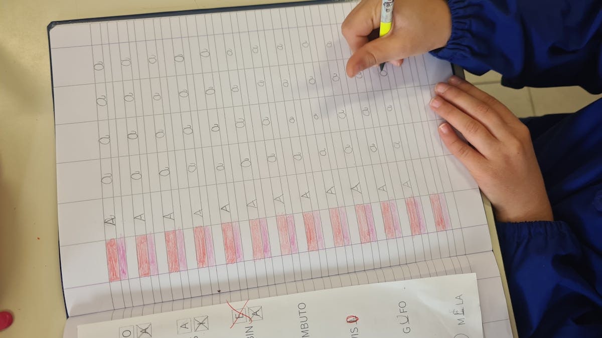A child in a blue sleeve writes letters in a notebook for educational practice.